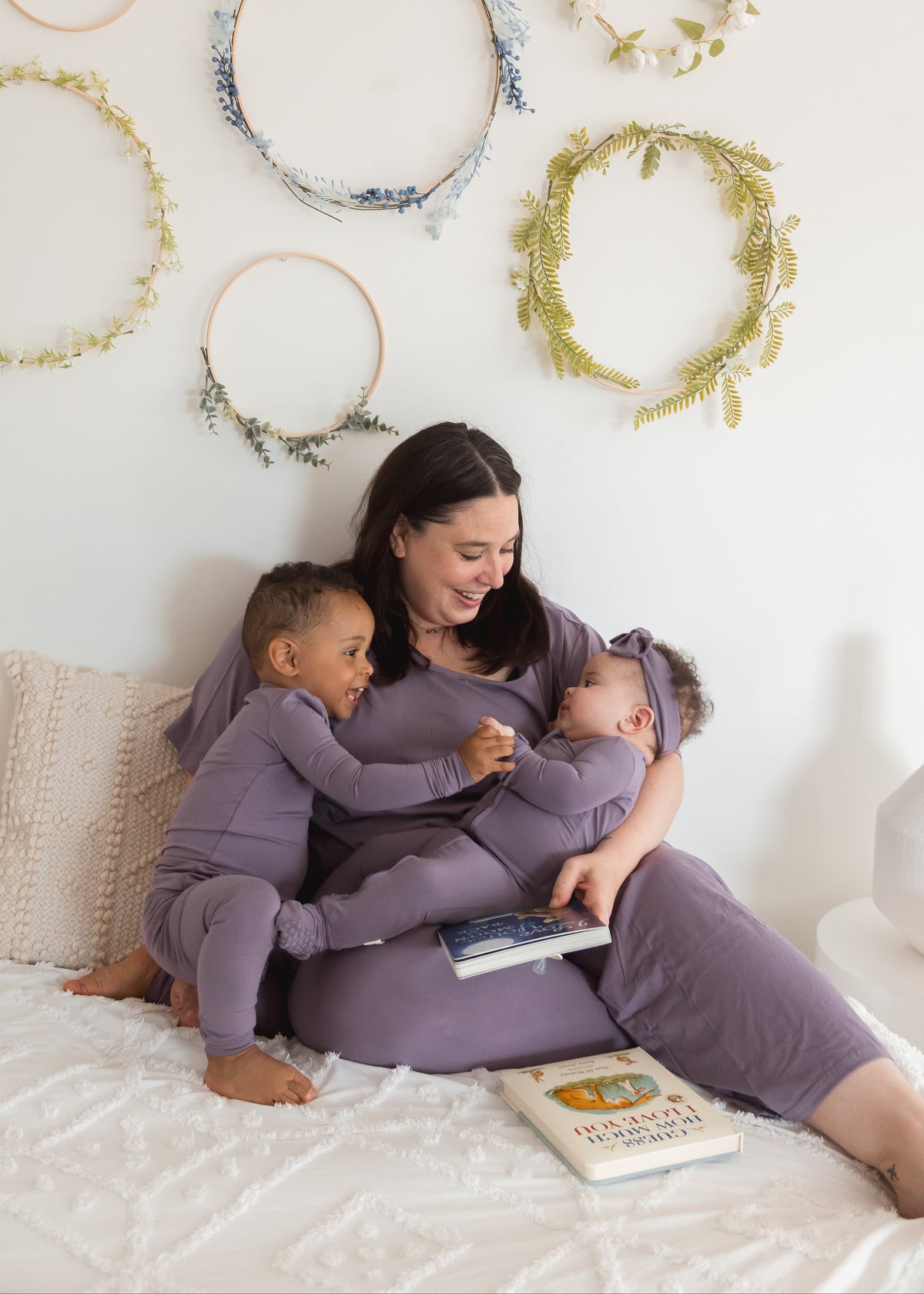 A woman and her two children wearing matching pajamas sit together on a white bed, with decorative floral hoops hanging on the wall behind them.