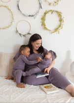 A woman and her two children wearing matching pajamas sit together on a white bed, with decorative floral hoops hanging on the wall behind them.