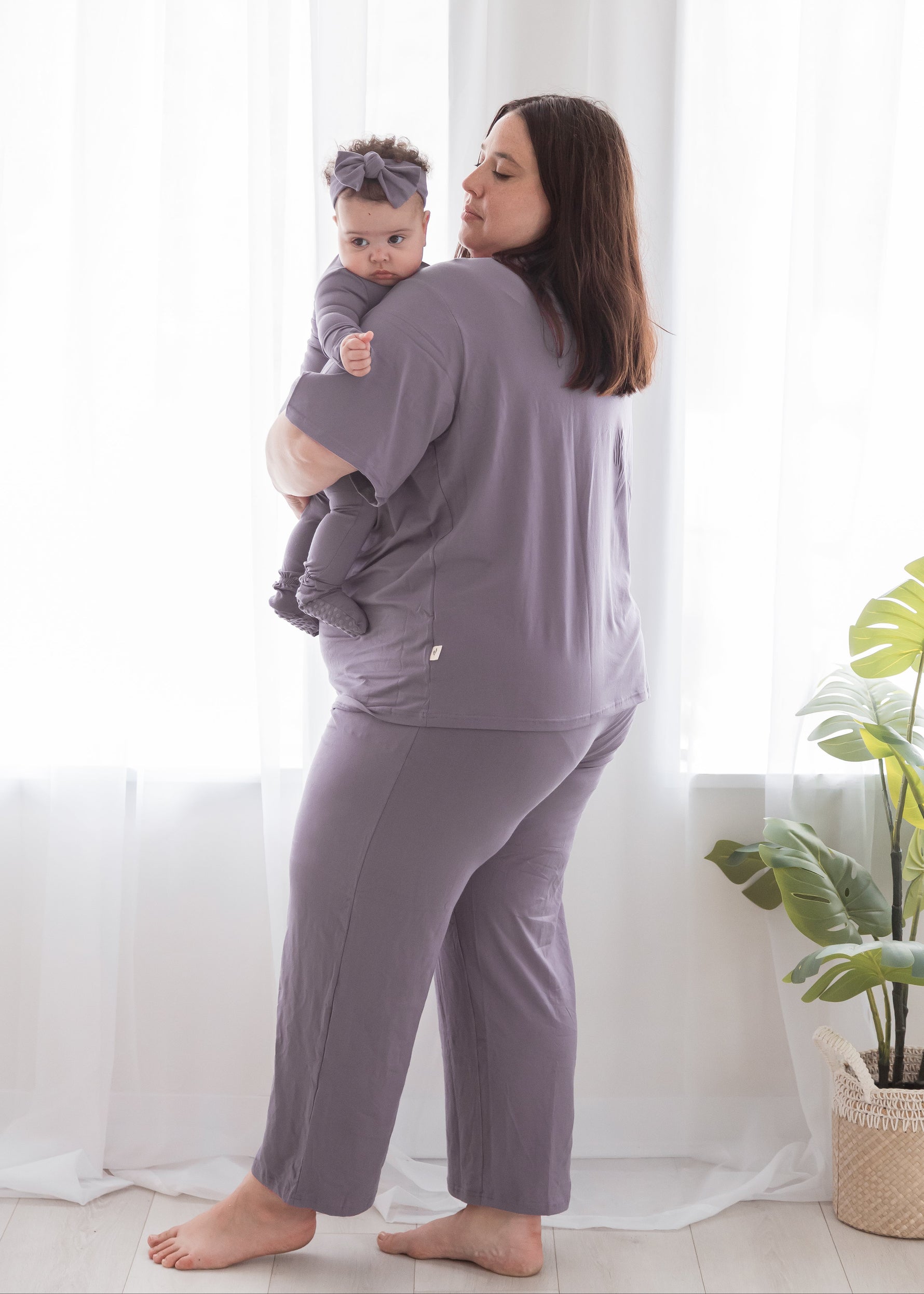 Side view of a mother holding her baby over her shoulder, both wearing matching Tencel pajamas in dusty purple; baby is in a zippered footie sleeper with a matching bow headband.