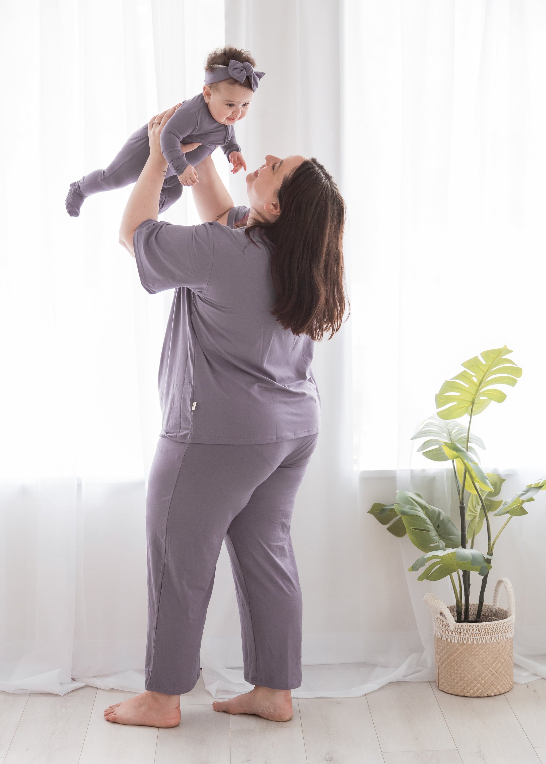 A mom and her baby wearing matching dusty purple loungewear set, including a short-sleeve top and pants, with the mom holding the baby up in a bright room with white curtains and a potted plant.