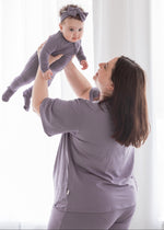 Happy mother lifting her baby in the air, both wearing matching dusty purple Tencel sleepwear, including a women's short-sleeve lounge set and a baby footie romper.