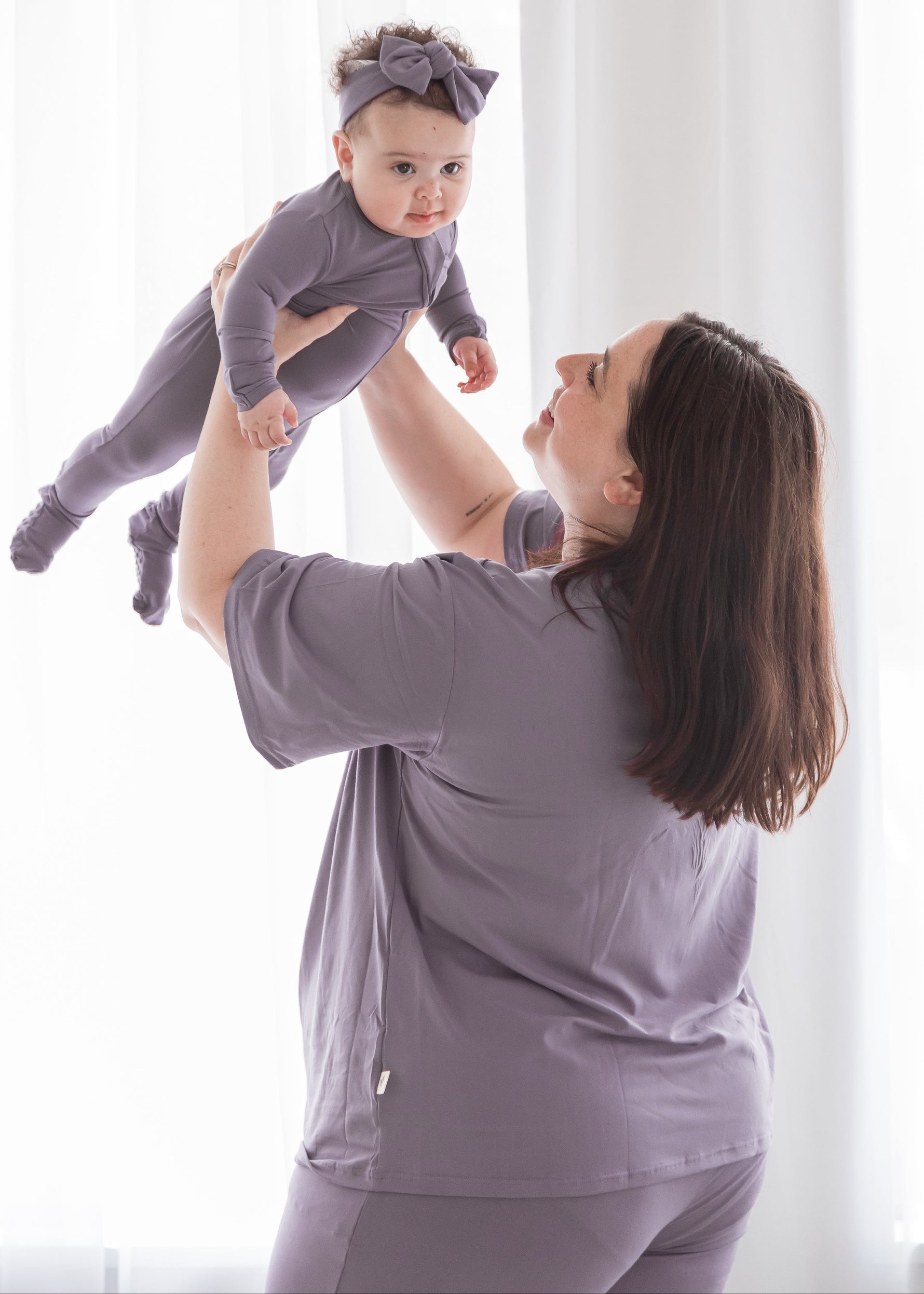 Happy mother lifting her baby in the air, both wearing matching dusty purple Tencel sleepwear, including a women's short-sleeve lounge set and a baby footie romper.