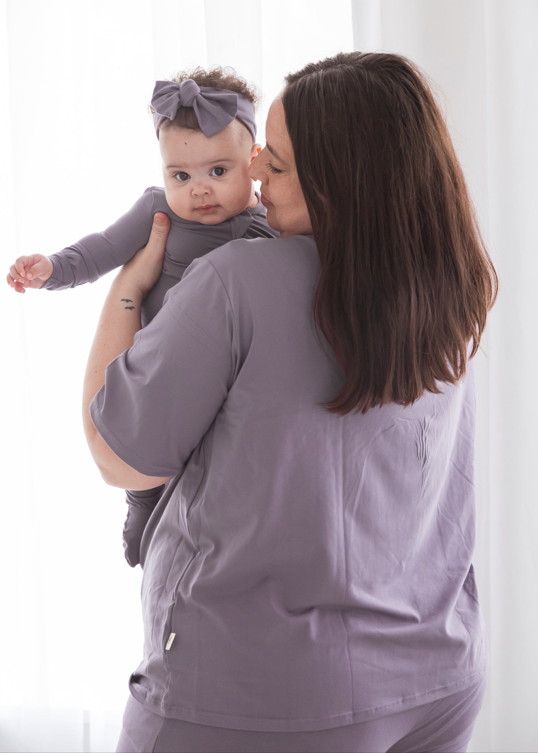 A mother from behind holding her baby over her shoulder; the mother wears matching Wild Mulberry purple loungewear and the baby wears a matching sleeper and headband.
