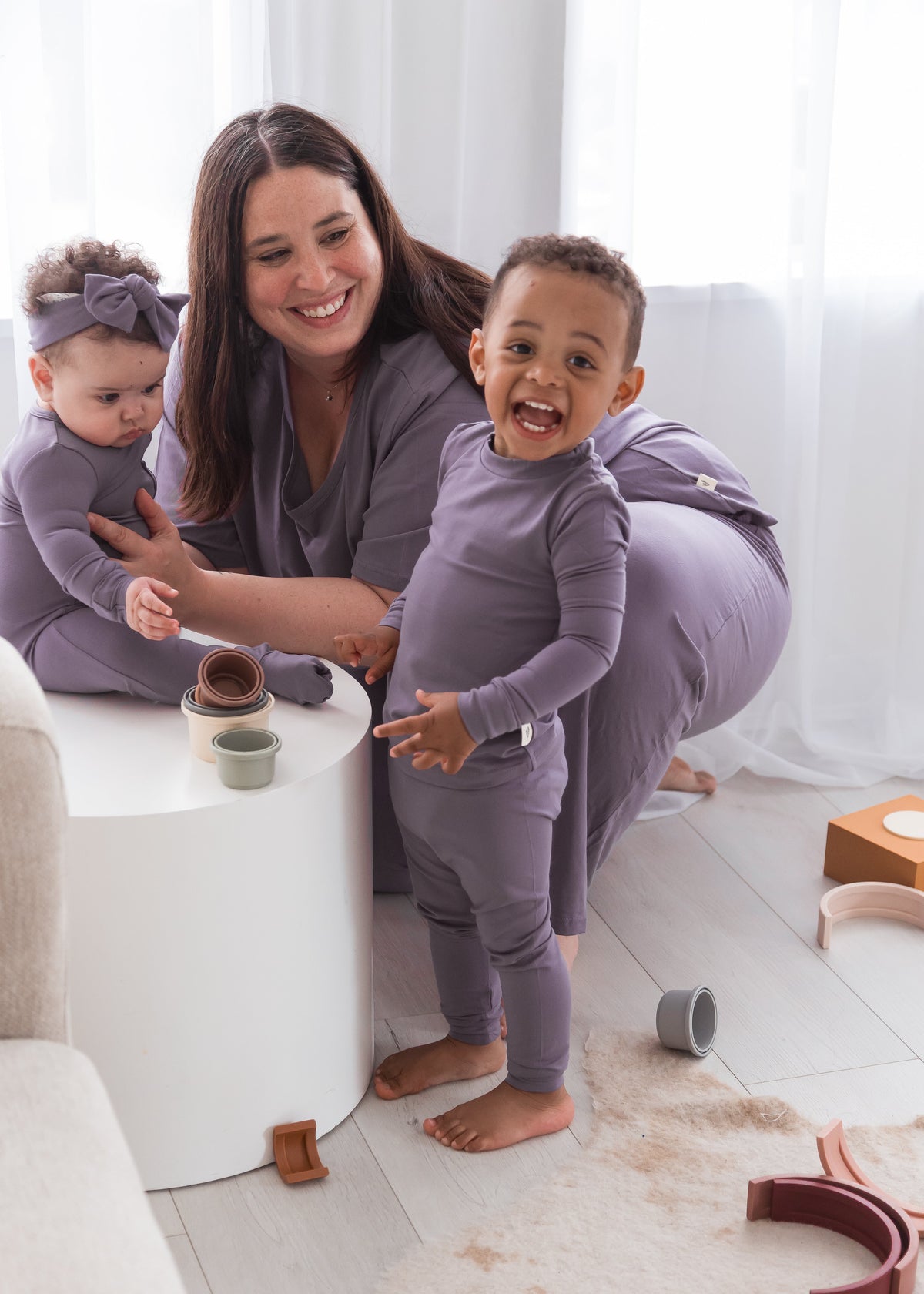 A smiling mother sitting with her baby and toddler in a bright room, all wearing matching Wild Mulberry purple TENCEL pajamas while playing with wooden toys.