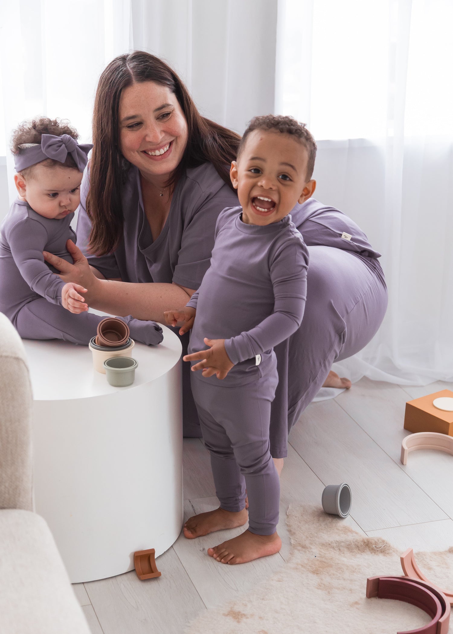 A smiling mother sitting with her baby and toddler in a bright room, all wearing matching Wild Mulberry purple TENCEL pajamas while playing with wooden toys.