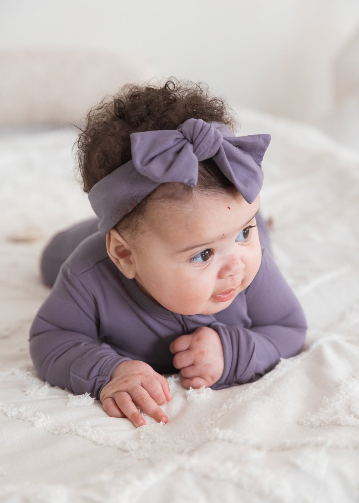 A baby lying on a white textured blanket wearing a Milana Label sleeper and matching dusty purple bow headband.