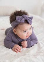 A baby lying on a white textured blanket wearing a Milana Label sleeper and matching dusty purple bow headband.