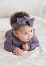A baby lying on a white textured blanket wearing a Milana Label sleeper and matching dusty purple bow headband.