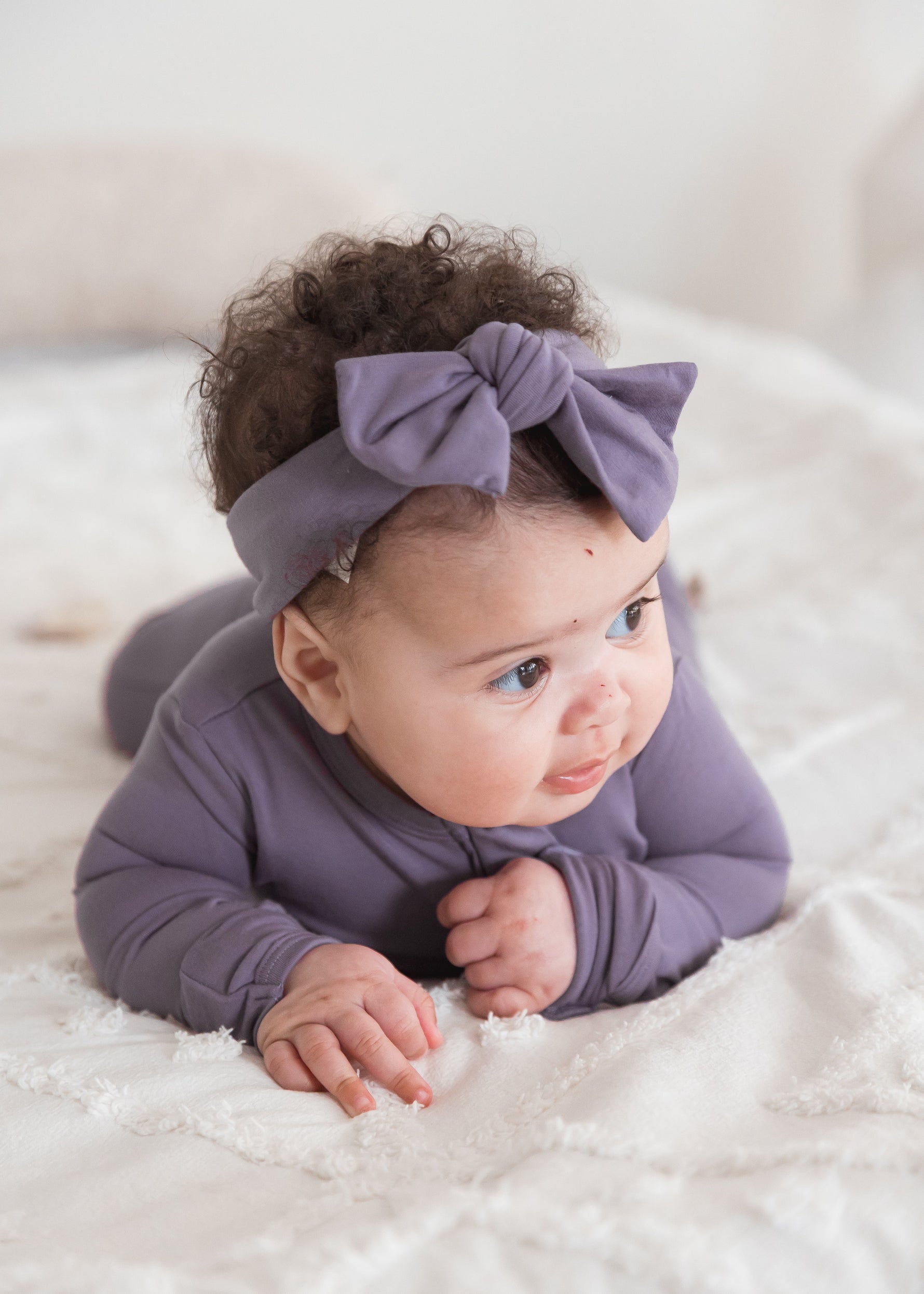 A baby lying on a white textured blanket wearing a Milana Label sleeper and matching dusty purple bow headband.