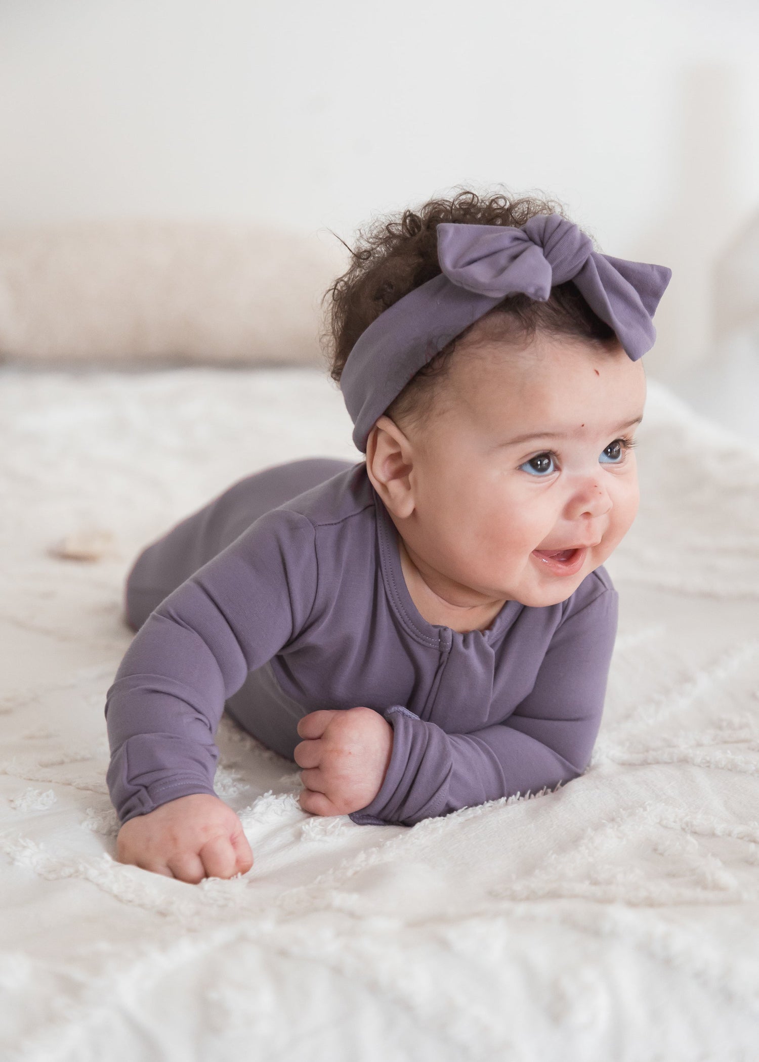 A baby wearing a matching TENCEL footie sleeper and bow headband, lying on a white textured blanket.