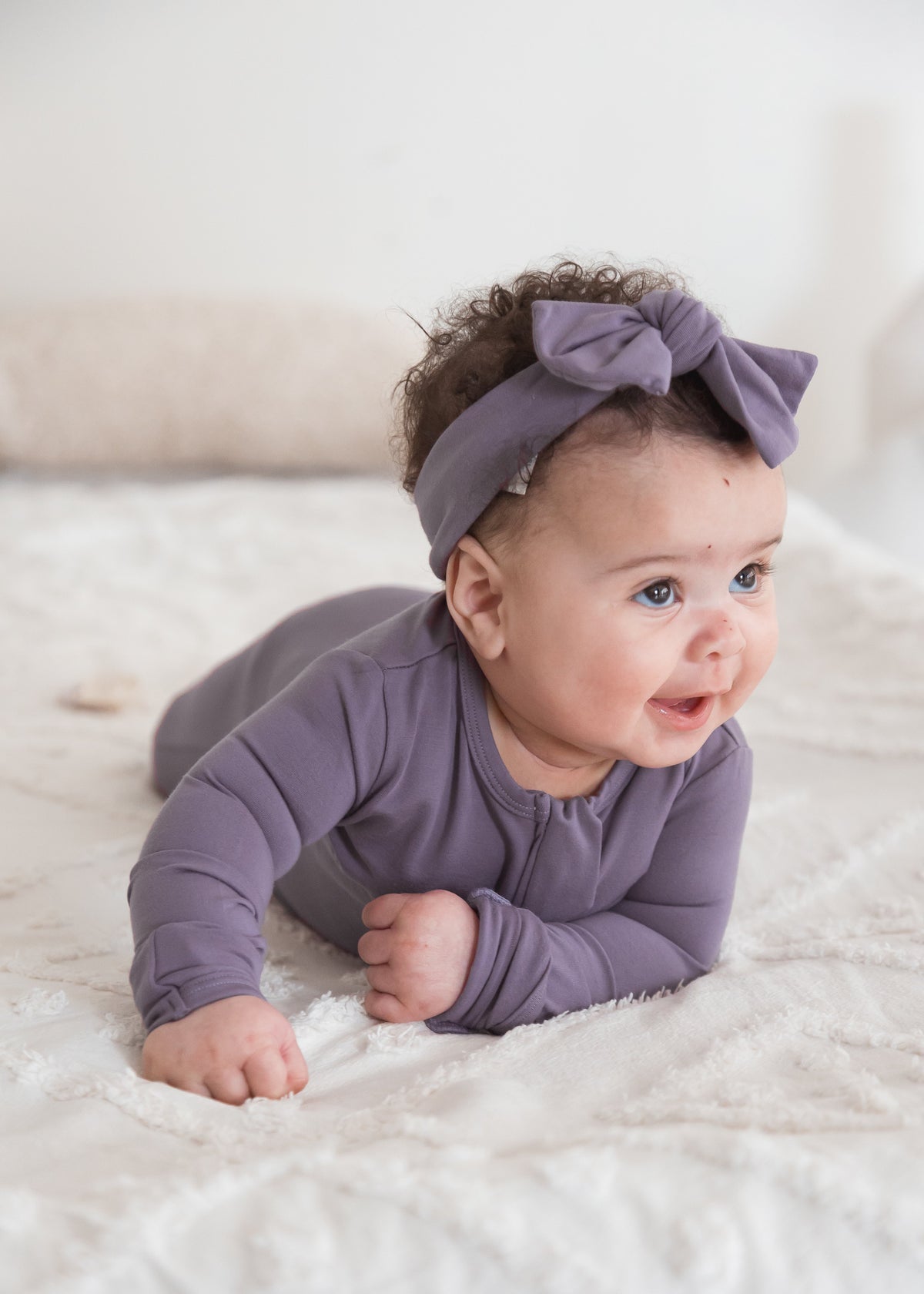A baby wearing a matching TENCEL footie sleeper and bow headband, lying on a white textured blanket.