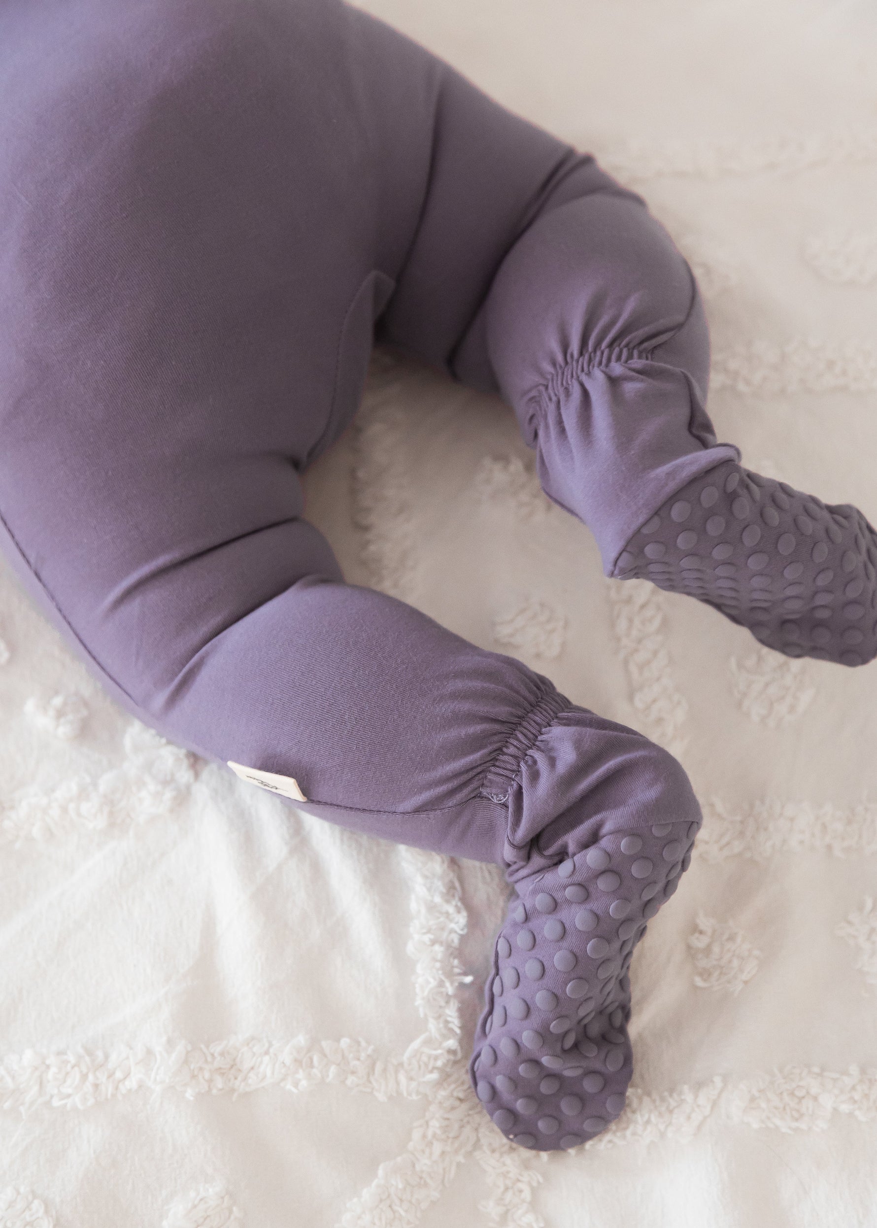 Detailed close-up of a baby's legs and feet wearing a Wild Mulberry purple sleeper, highlighting the patterned grip soles on the footies and elastic ankle backing.