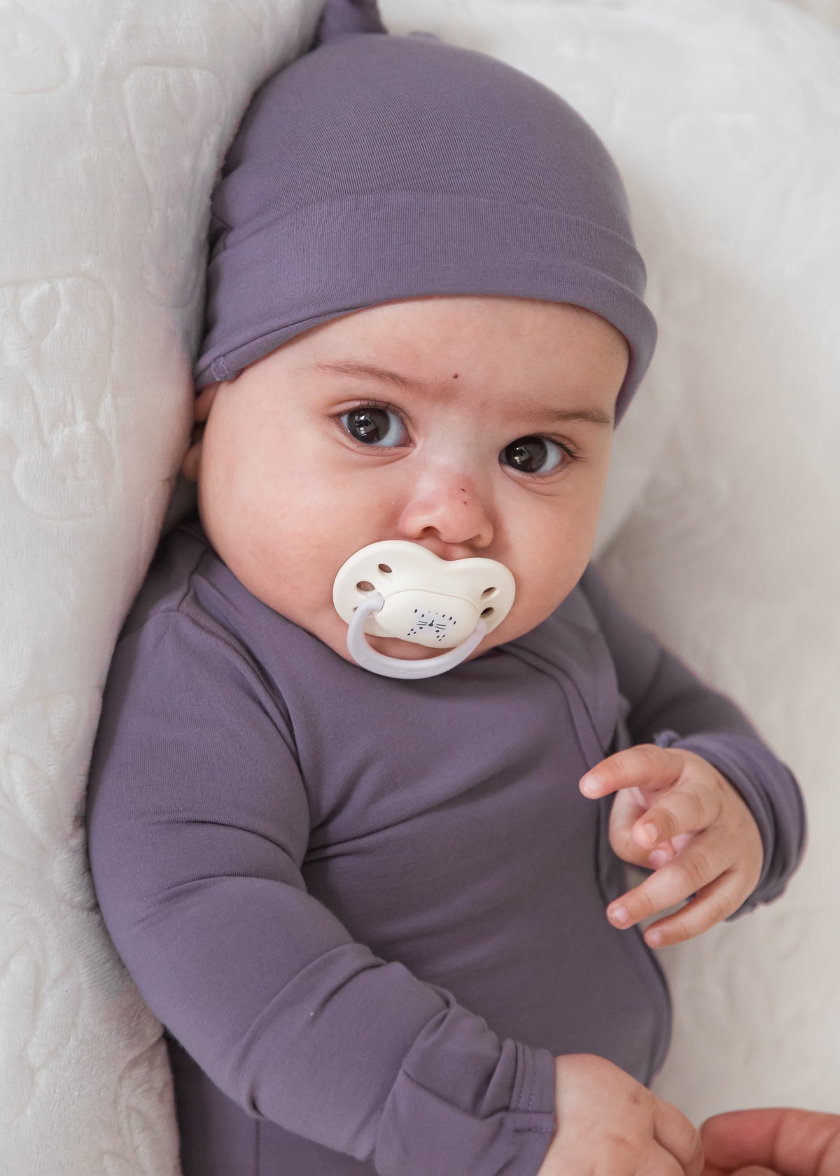 A baby wearing a matching dusty purple long-sleeve sleeper and hat lies on a white quilted mattress, with a white pacifier in their mouth and an adult's hand adjusting the bodysuit.