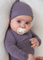 A baby wearing a matching dusty purple long-sleeve sleeper and hat lies on a white quilted mattress, with a white pacifier in their mouth and an adult's hand adjusting the bodysuit.
