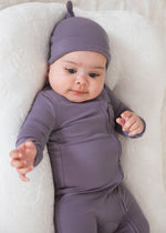 A baby lying comfortably on a white lounger pillow, dressed in a Wild Mulberry purple long-sleeve sleeper and matching knotted beanie hat.
