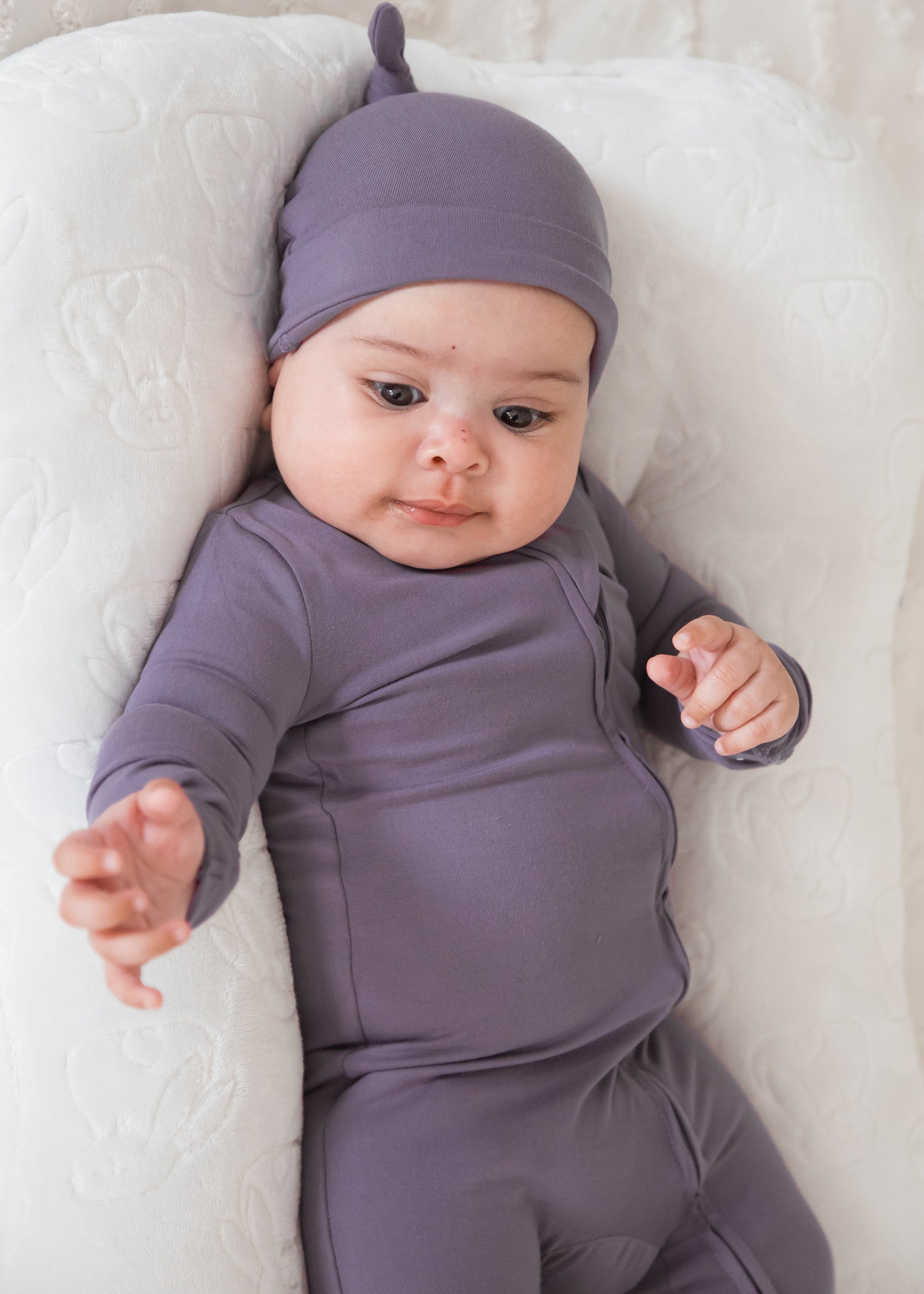A baby lying comfortably on a white lounger pillow, dressed in a Wild Mulberry purple long-sleeve sleeper and matching knotted beanie hat.