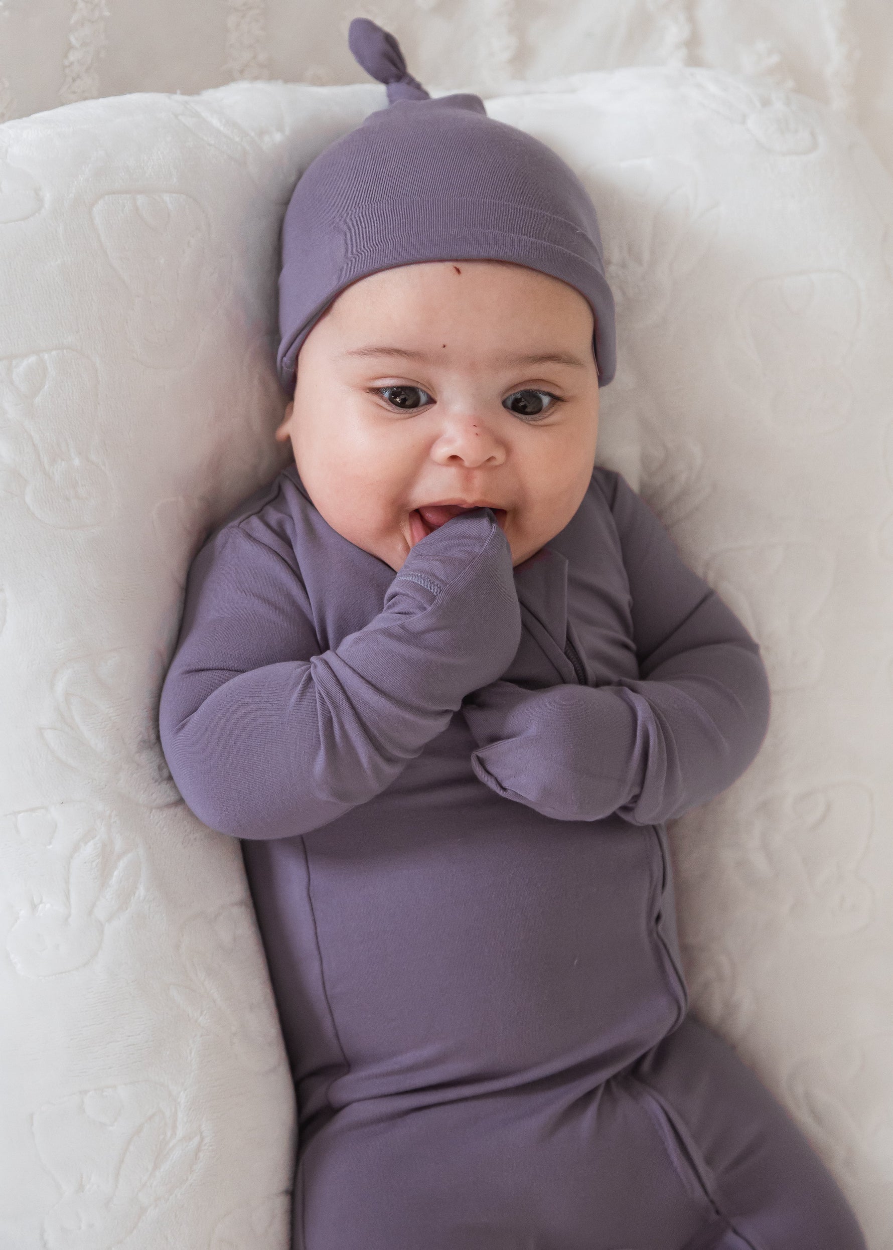 Close-up of a baby lying on a white lounger cushion, wearing a Wild Mulberry purple sleeper with fold-over mittens and a matching knotted beanie hat.