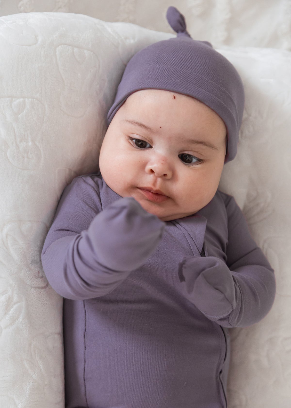 A baby wearing a matching Milana Label long-sleeve bodysuit and knot bonnet, lying on a white quilted mattress.