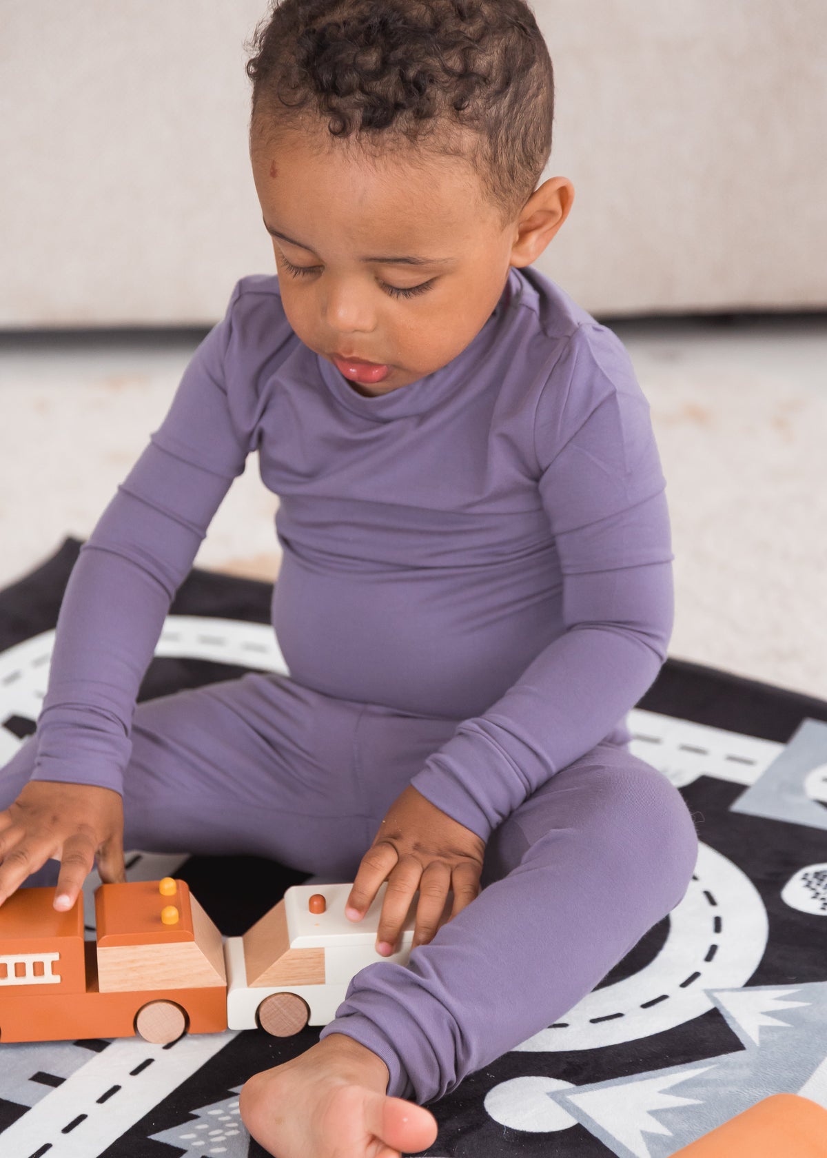 A toddler wearing a matching long-sleeve one-piece pajama set in dusty purple, sitting on a black and white play rug and playing with a wooden toy truck.