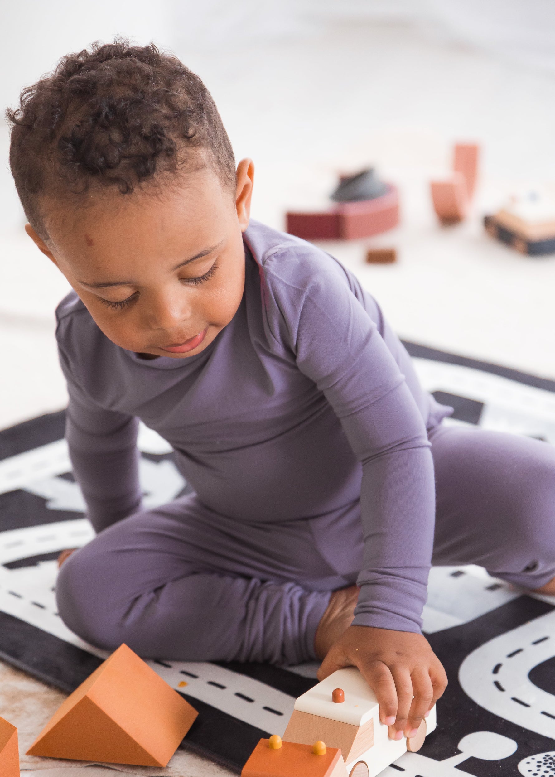 Close-up of a toddler sitting on a play mat, focused on playing with wooden block toys while wearing a purple Wild Mulberry TENCEL long-sleeve pajama set.