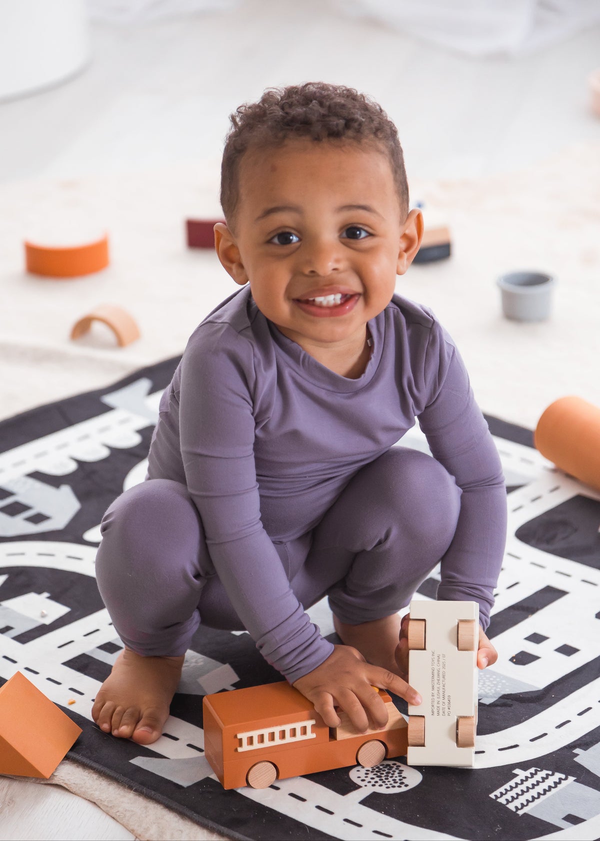 A smiling toddler crouching on a black and white road play mat, wearing a Wild Mulberry purple TENCEL pajama set and playing with a wooden toy train.