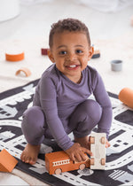 A smiling toddler crouching on a black and white road play mat, wearing a Wild Mulberry purple TENCEL pajama set and playing with a wooden toy train.