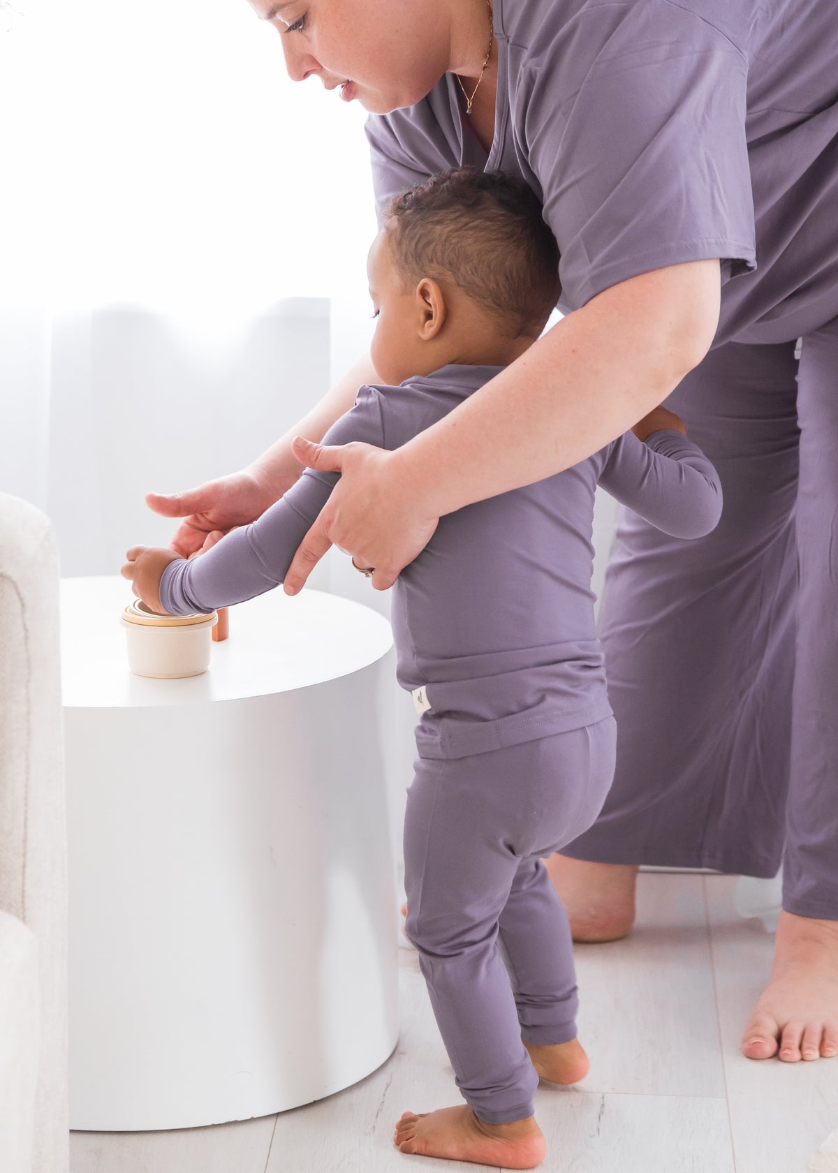 A toddler and an adult both wearing matching lavender-purple pajamas, standing barefoot in a bright room next to a white side table.