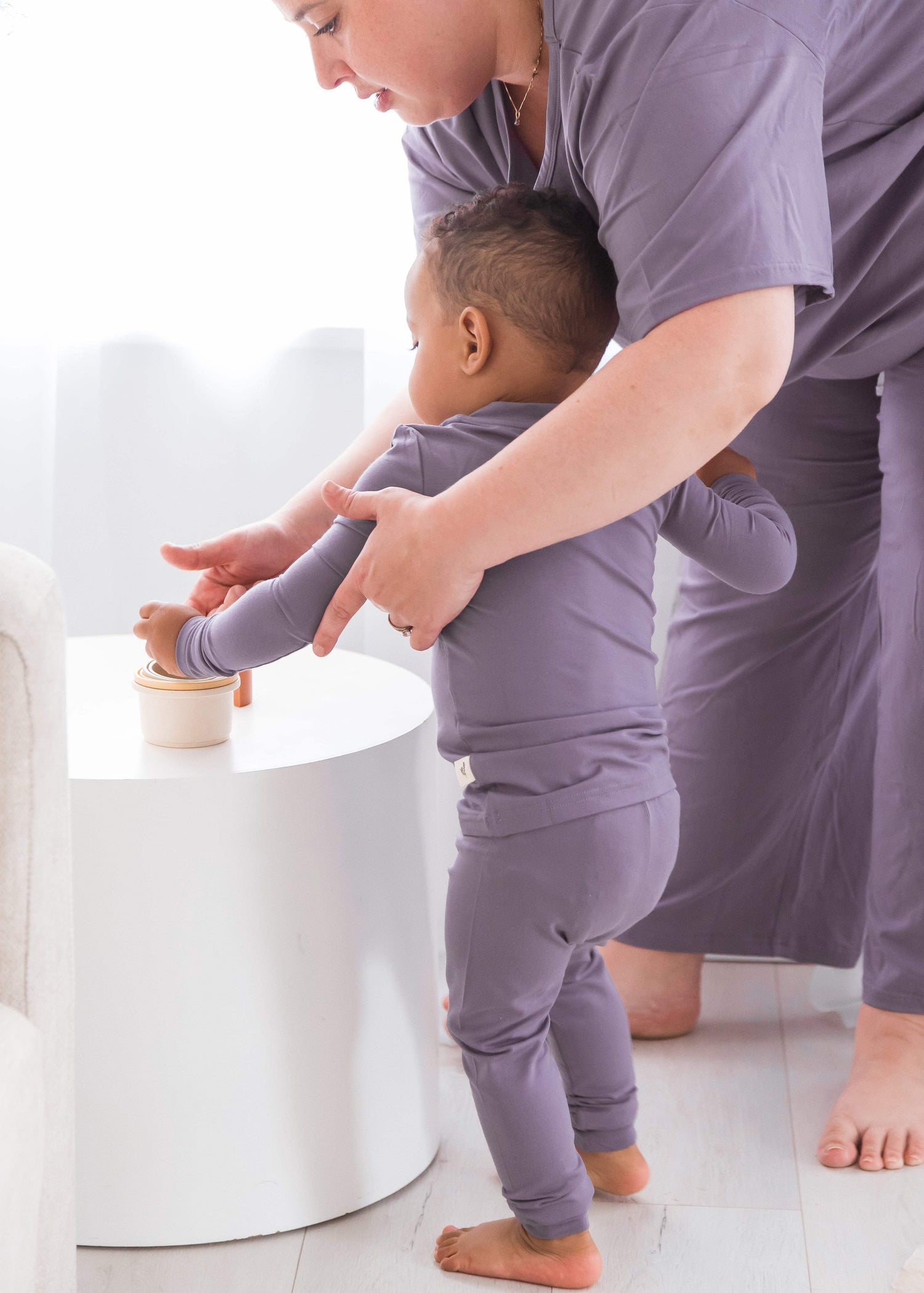 A toddler and an adult both wearing matching lavender-purple pajamas, standing barefoot in a bright room next to a white side table.