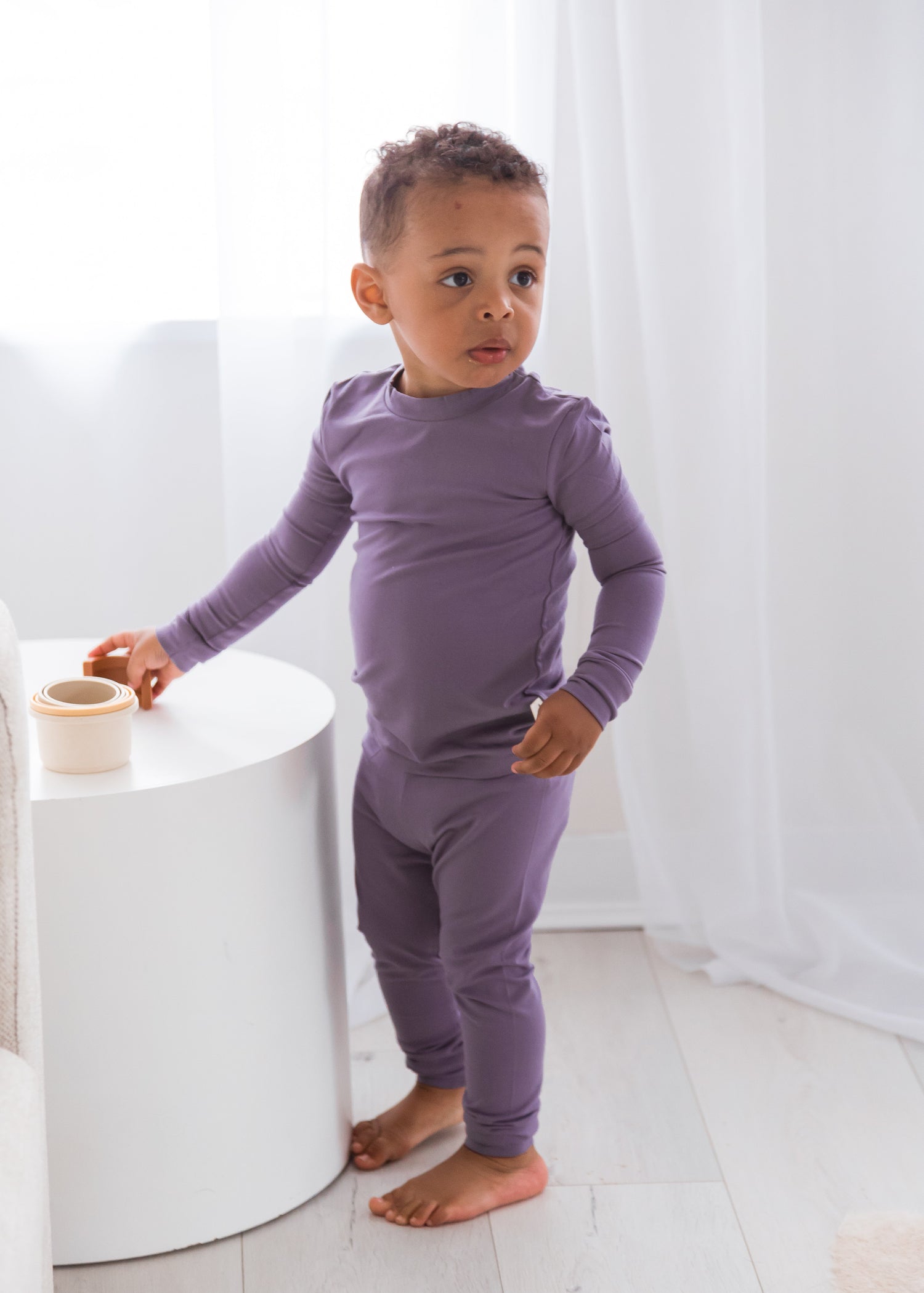 A toddler standing next to a white table in a brightly lit room, dressed in a solid purple Wild Mulberry TENCEL two-piece pajama set.