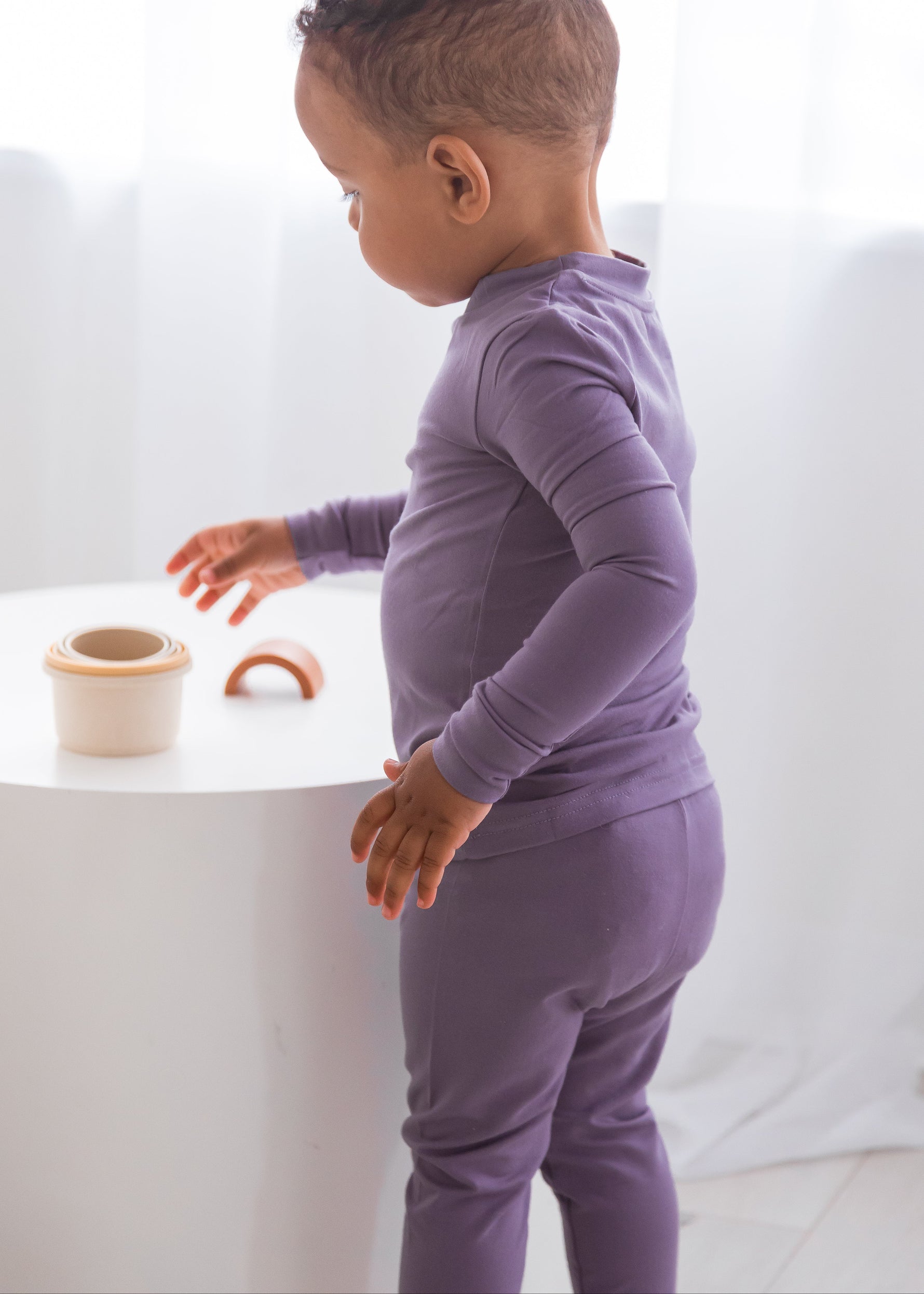 Side view of a toddler standing by a white cylindrical table, wearing a snug-fit Wild Mulberry purple TENCEL long-sleeve pajama set.