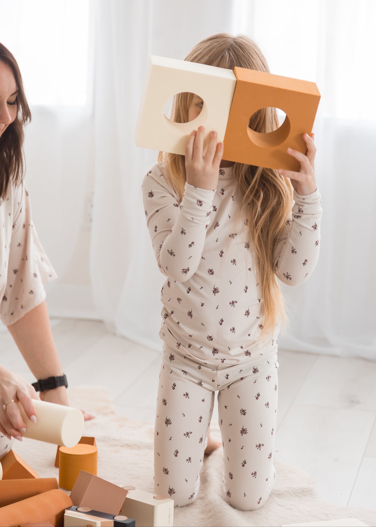 A playful shot of a young girl in a birch floral Tencel PJ set holding cream and orange foam building blocks up to her face like glasses, with her mother sitting beside her.