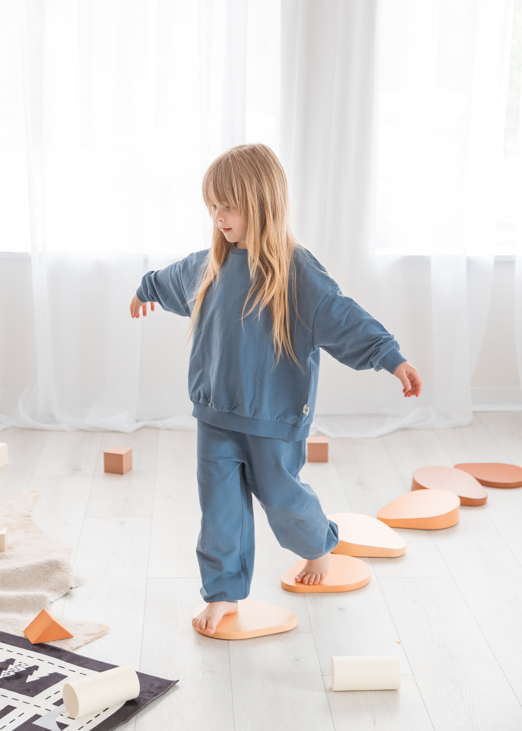 A young girl with long blonde hair steps across wooden balancing blocks in a sunlit playroom, wearing a matching Nordic blue slate-toned organic cotton tracksuit.
