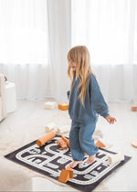 A young girl with long blonde hair stands barefoot on a black and white road-map style rug in a bright, modern room. She is wearing a matching blue sweatshirt and sweatpants set, surrounded by wooden blocks.