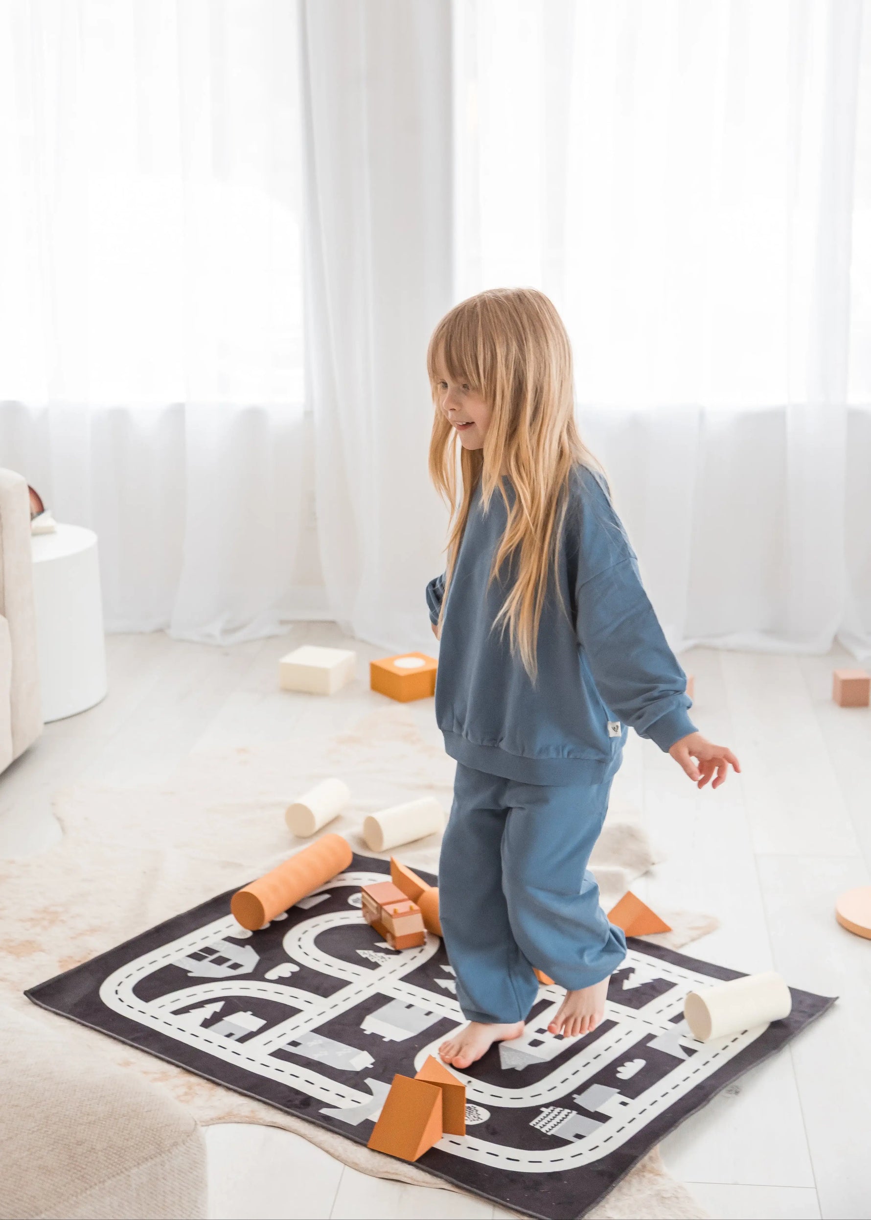 A young girl with long blonde hair stands barefoot on a black and white road-map style rug in a bright, modern room. She is wearing a matching blue sweatshirt and sweatpants set, surrounded by wooden blocks.