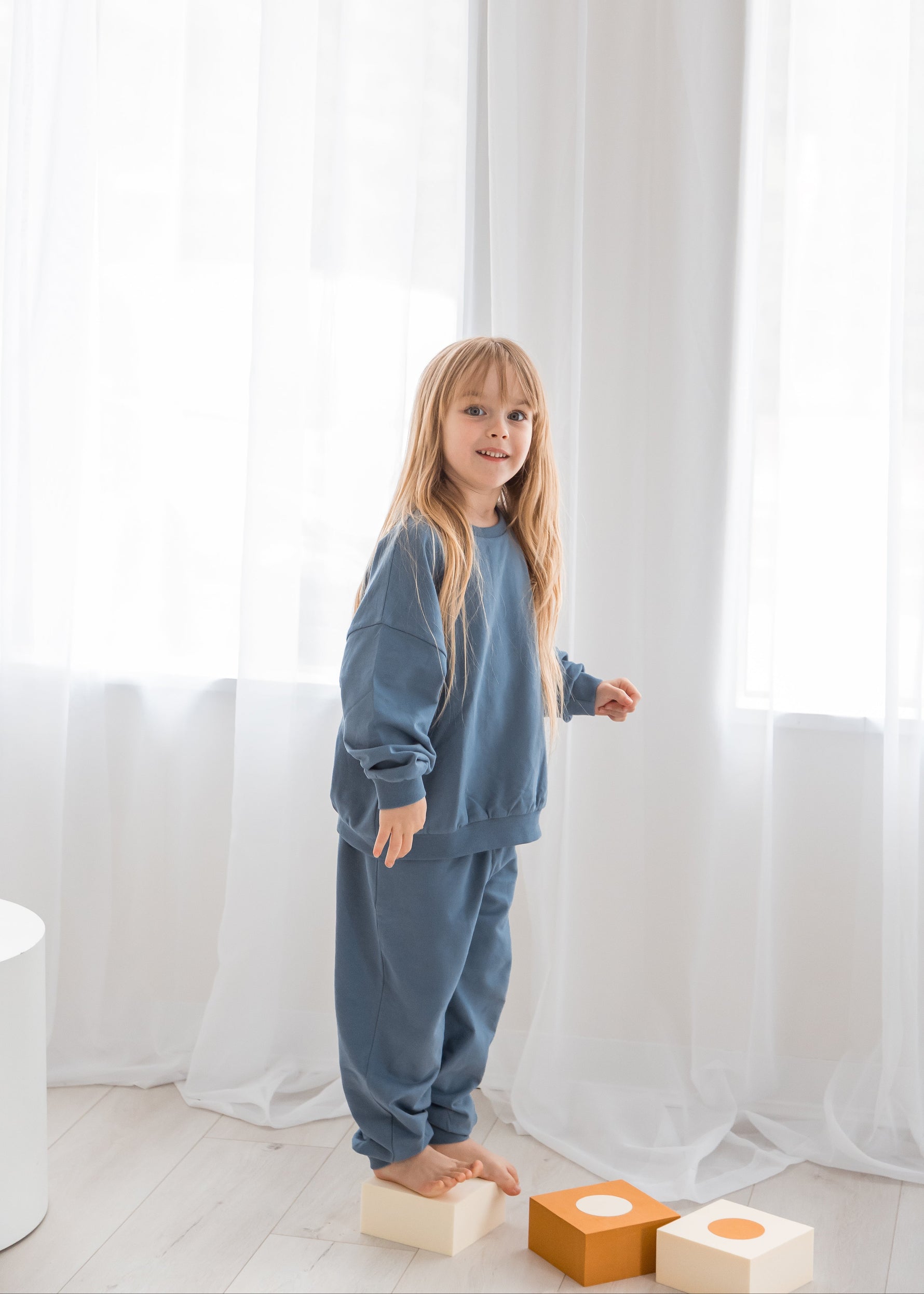 A young girl in a matching Nordic blue organic cotton two-piece sweater and pants set stands balanced on a wooden toy block in a bright, neutral-toned kids' playroom.