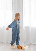 Side profile view of a young girl wearing a cozy Nordic blue organic cotton jogger set as she balances on top of wooden stepping blocks near bright window curtains.