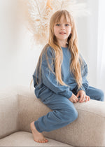 A young girl smiles while sitting on a neutral-colored fabric sofa, dressed in a cozy, matching Nordic blue organic cotton sweatshirt and jogger pants set.