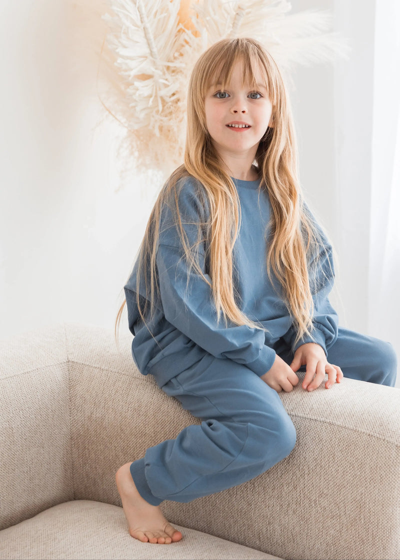 A young girl smiles while sitting on a neutral-colored fabric sofa, dressed in a cozy, matching Nordic blue organic cotton sweatshirt and jogger pants set.