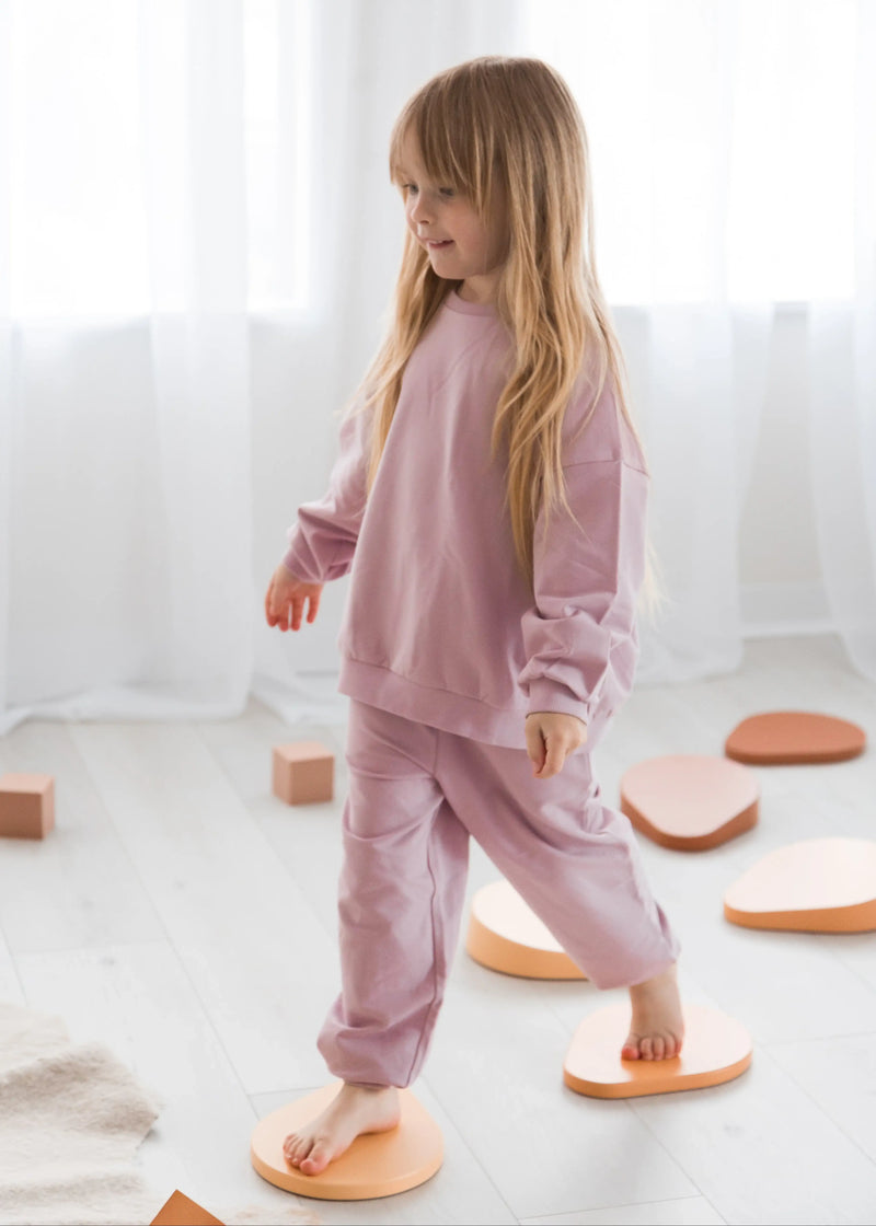 Side profile view of a young girl wearing a cozy, pale orchid organic cotton loungewear set as she balances across wooden stepping stones near bright window curtains.
