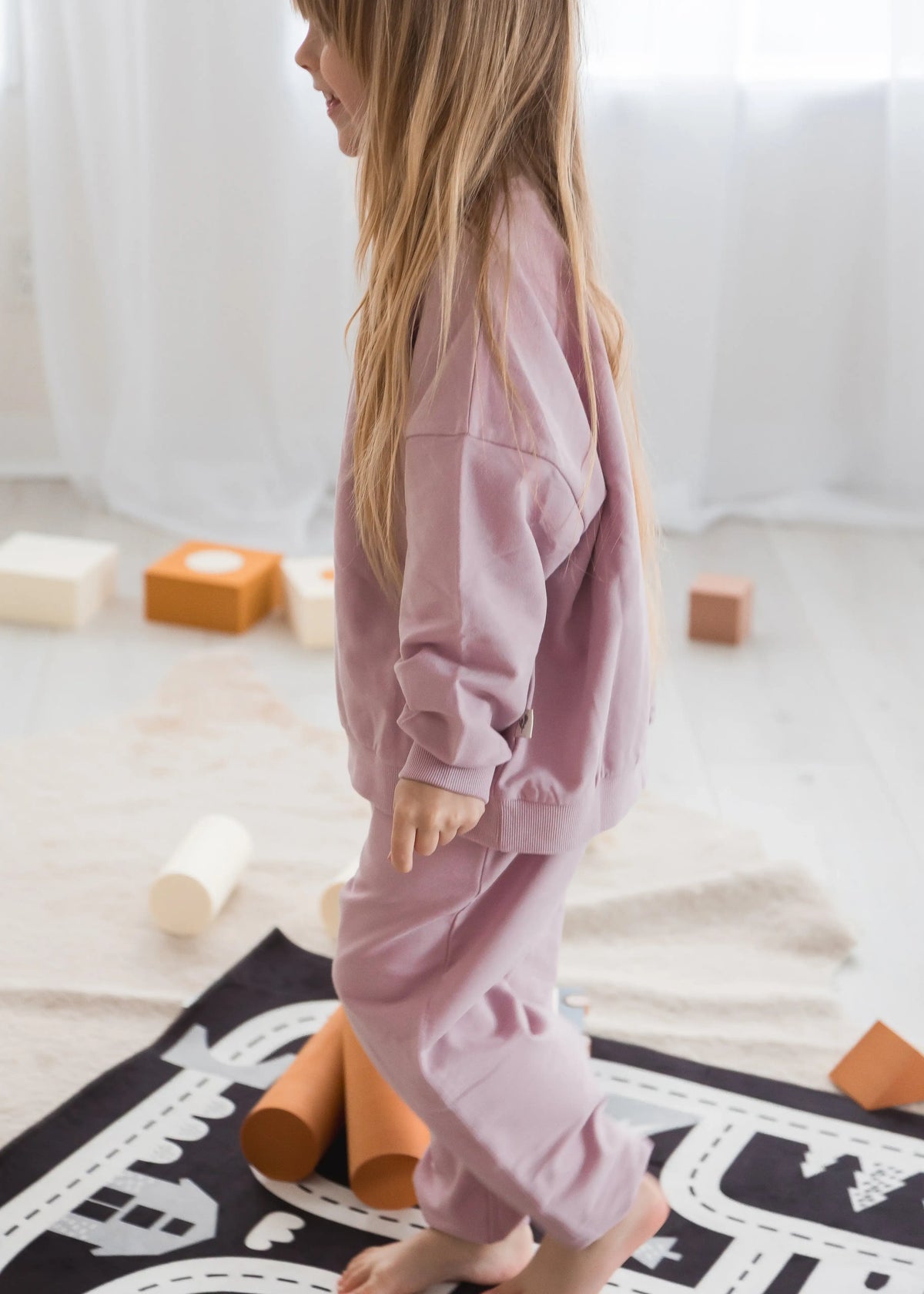 A young girl with long blonde hair is shown from the side, wearing dusty pink sweatshirt and jogger pants set. She is standing barefoot on a black and white play mat in a bright, neutral room with wooden blocks and foam rollers.
