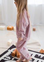 A young girl with long blonde hair is shown from the side, wearing dusty pink sweatshirt and jogger pants set. She is standing barefoot on a black and white play mat in a bright, neutral room with wooden blocks and foam rollers.