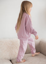 A young girl in a matching pale orchid muted pink organic cotton sweater and pants set steps along the cushions of a cream-colored sofa in a minimalist living room.