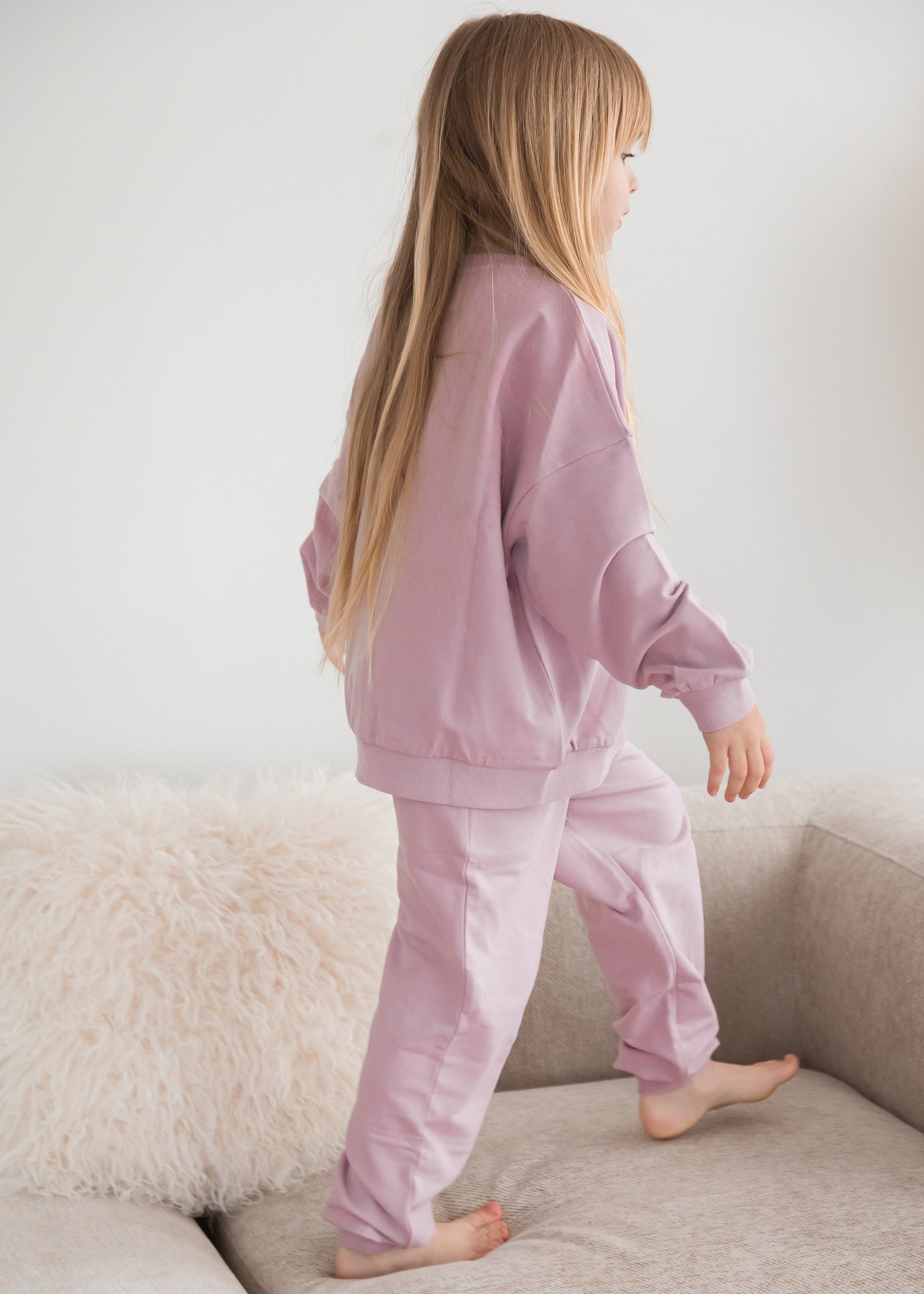 A young girl in a matching pale orchid muted pink organic cotton sweater and pants set steps along the cushions of a cream-colored sofa in a minimalist living room.