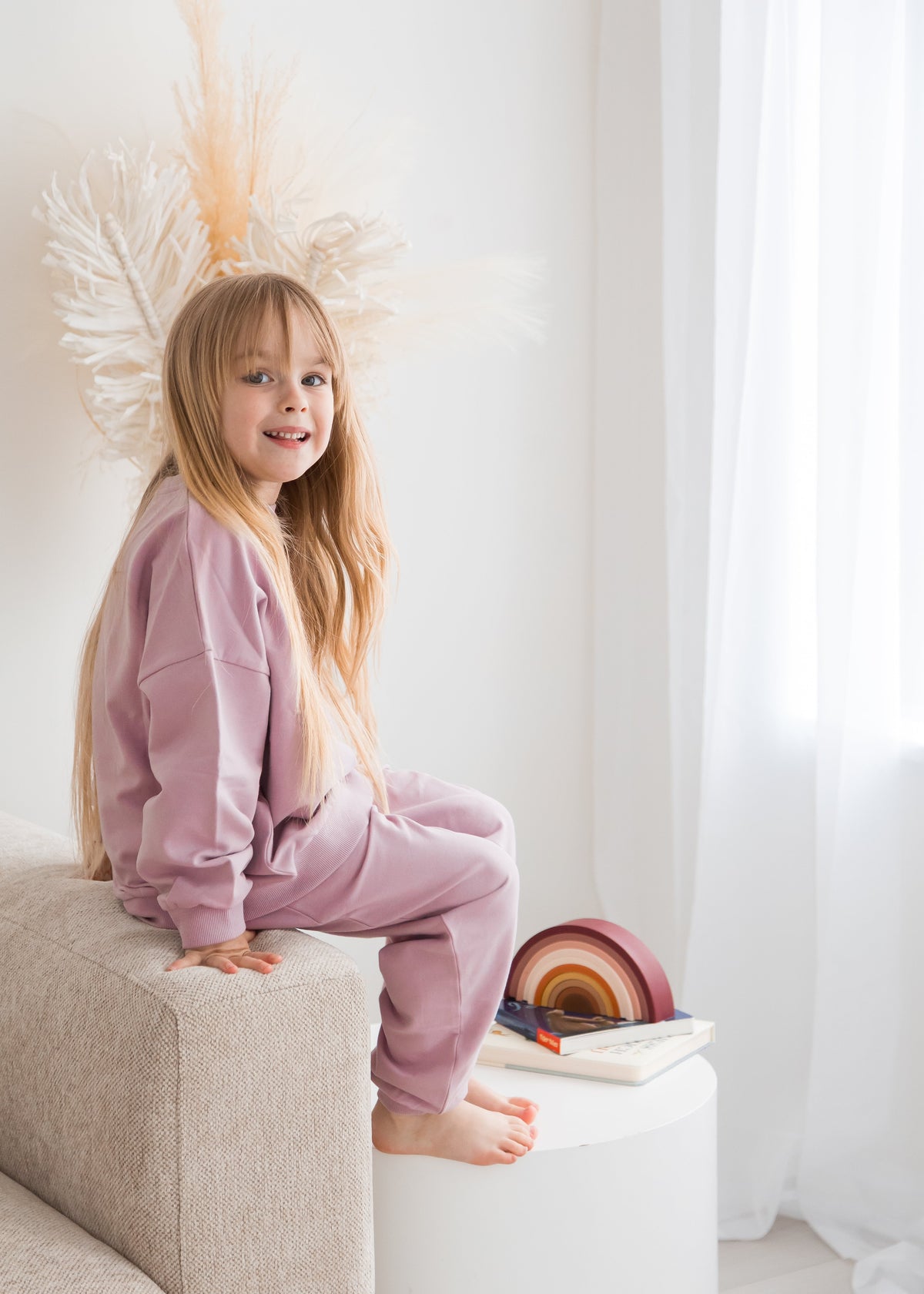 Full-length view of a young child sitting on the armrest of a neutral-toned sofa, wearing a matching pale orchid dusty pink organic cotton sweatshirt and jogger pants set.
