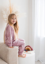 Full-length view of a young child sitting on the armrest of a neutral-toned sofa, wearing a matching pale orchid dusty pink organic cotton sweatshirt and jogger pants set.