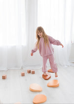 A young girl with long blonde hair steps across wooden stepping stones while wearing a loose-fit, muted pink organic cotton sweatshirt and matching pants in a bright, minimalist playroom.