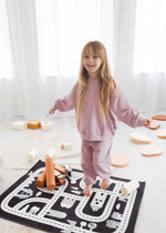 A smiling young girl stands on a monochrome road-patterned play mat in a sunlit room, wearing a cozy pale orchid organic cotton lounge set.