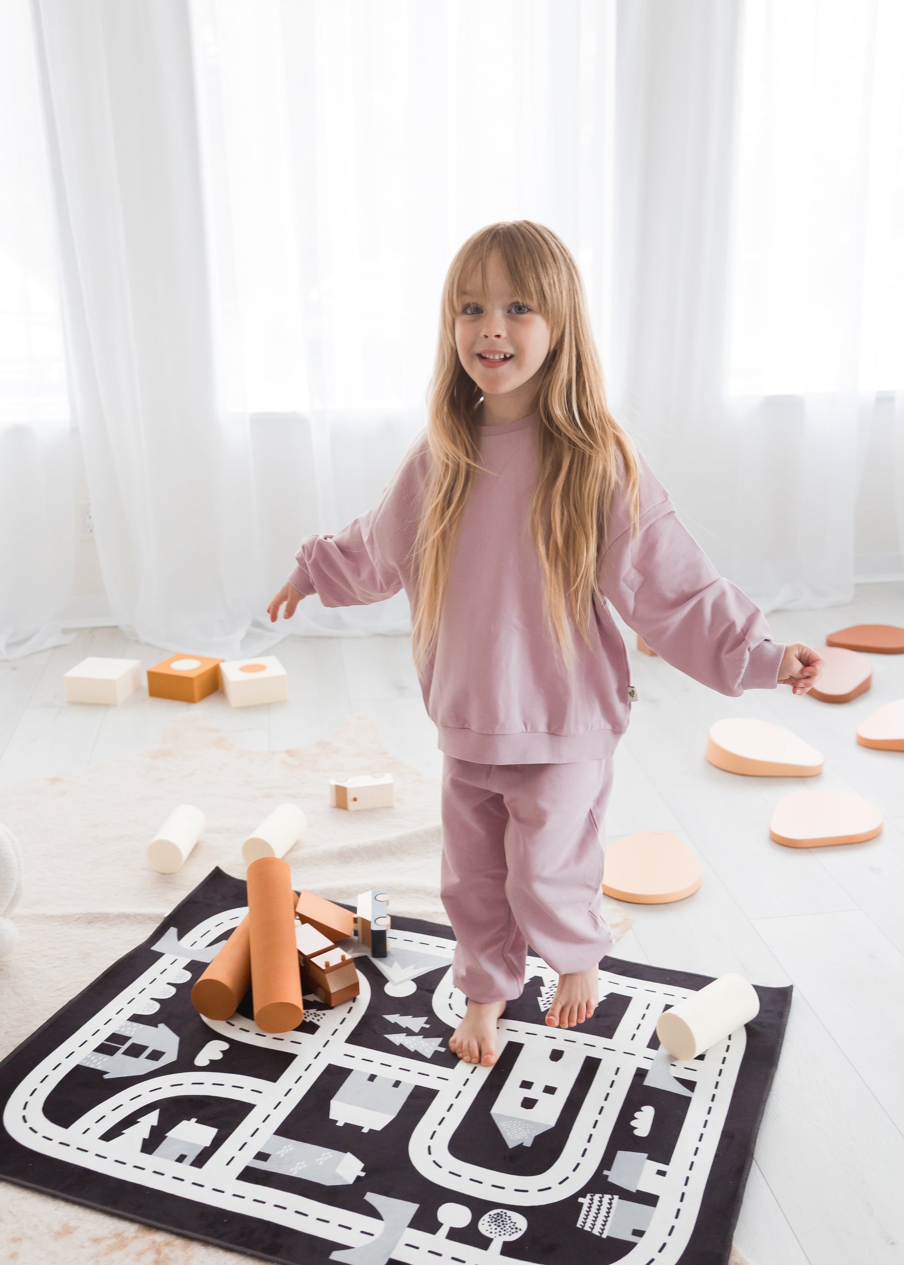 A smiling young girl stands on a monochrome road-patterned play mat in a sunlit room, wearing a cozy pale orchid organic cotton lounge set.