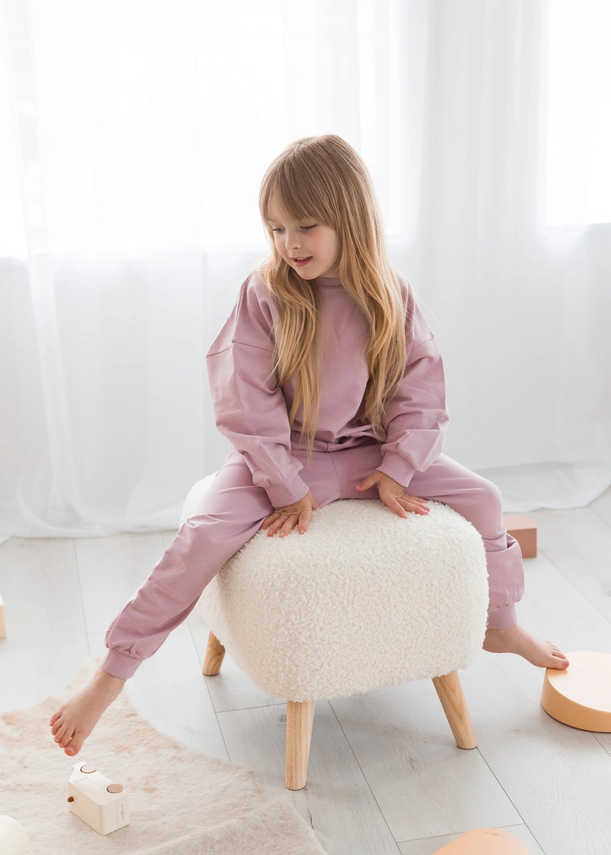 A young girl with long blonde hair sits barefoot on a white sheepskin ottoman, wearing an organic cotton jogger set in a bright, modern room with white curtains and light wood flooring.