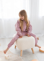 A young girl with long blonde hair sits barefoot on a white sheepskin ottoman, wearing an organic cotton jogger set in a bright, modern room with white curtains and light wood flooring.
